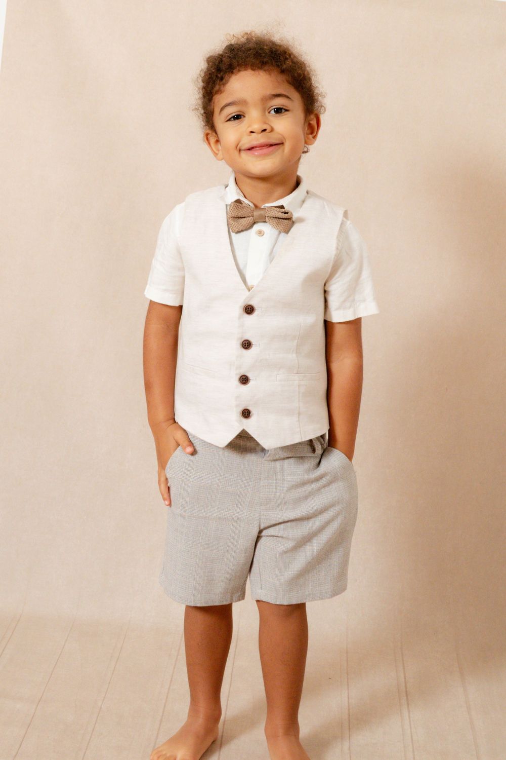 Young boy in a cream studio setting wearing tailored shorts with a waistcoat and bow tie.