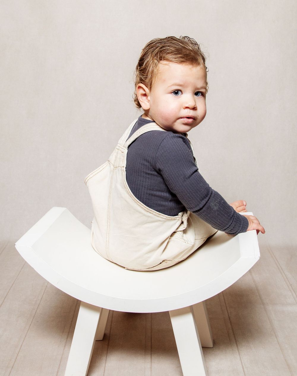 Cream studio setting of a young boy sitting on a white curved chair looking toward the camera.