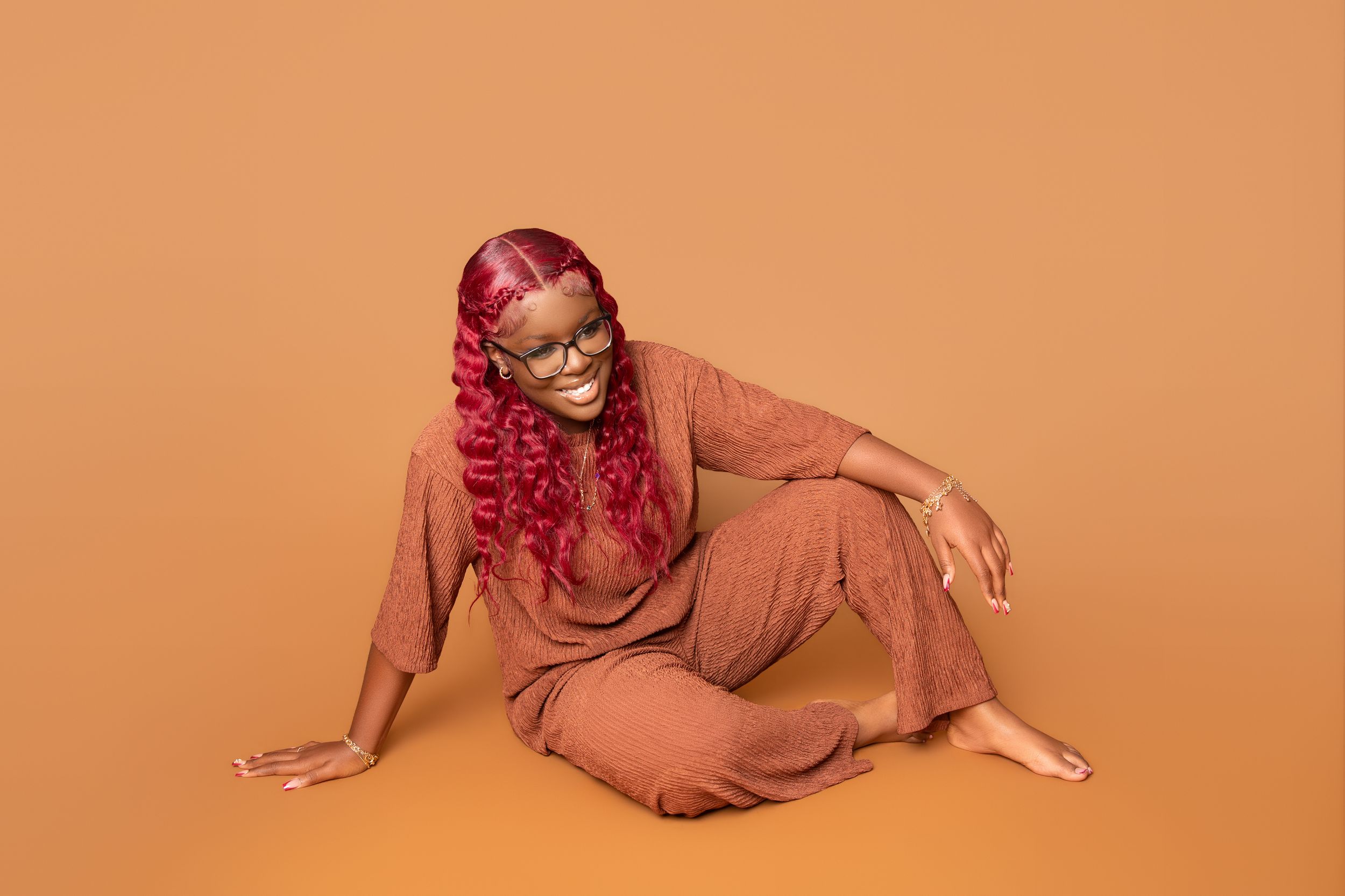 Person with vibrant red hair wearing a brown outfit poses on orange background in artistic studio setting.