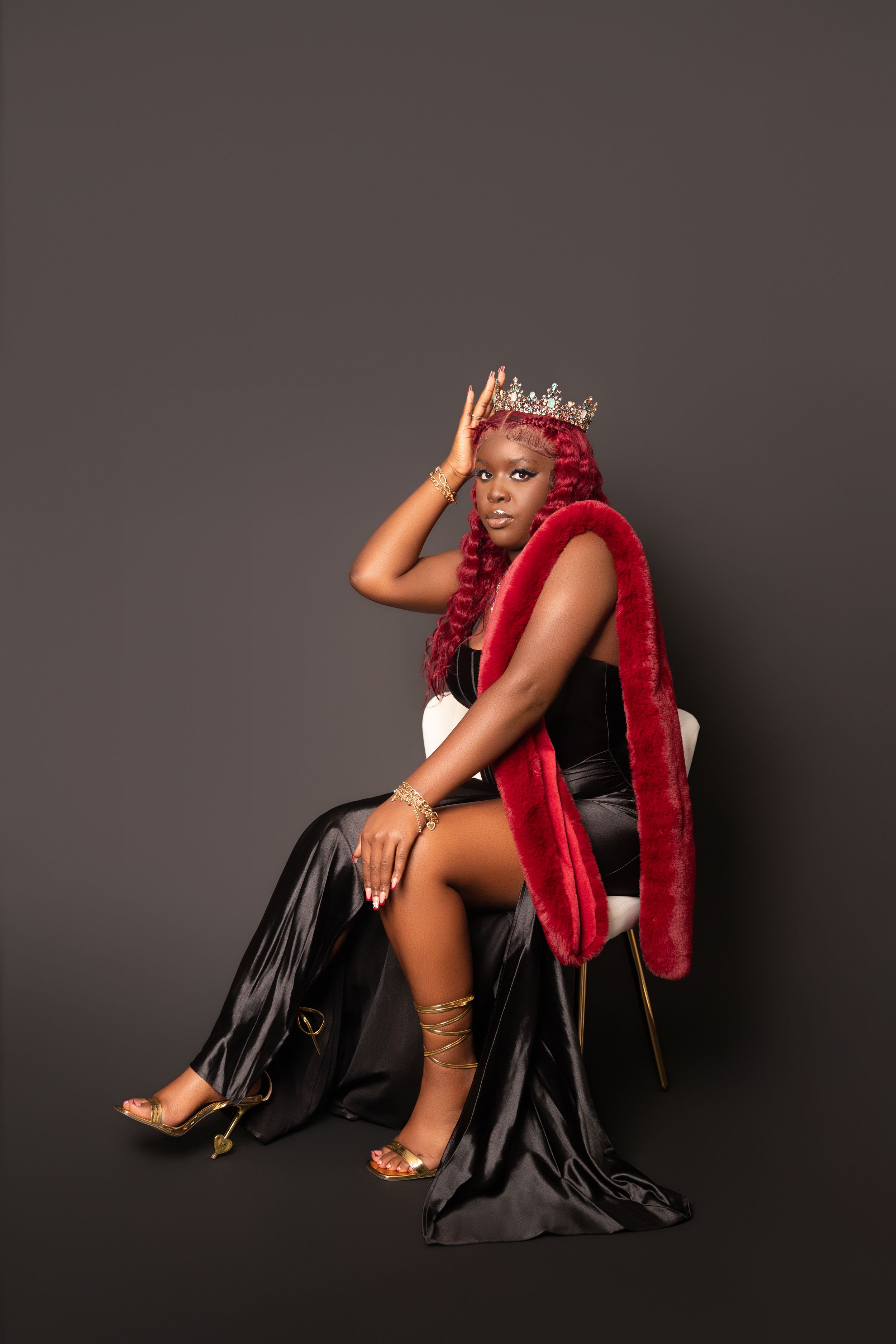 A glamorous fashion portrait featuring a black satin dress, bright red boa, and strappy gold sandals against a dark background.