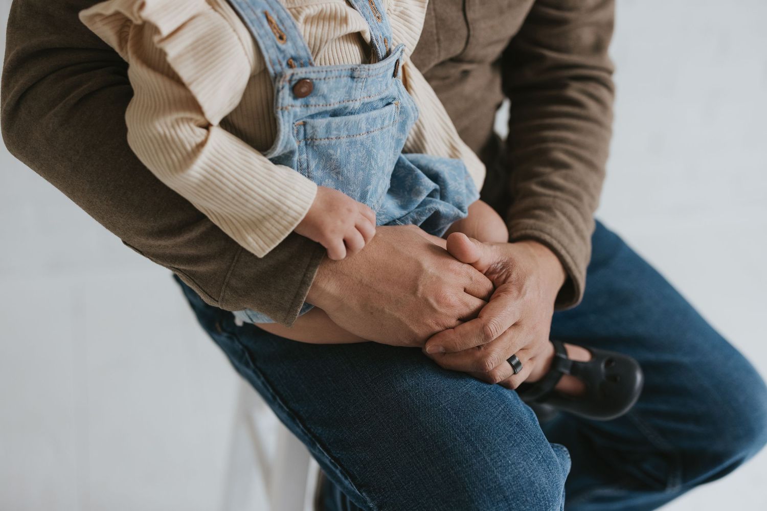 Person sitting in denim overalls with hands clasped together on knees, wearing a brown sweater underneath.