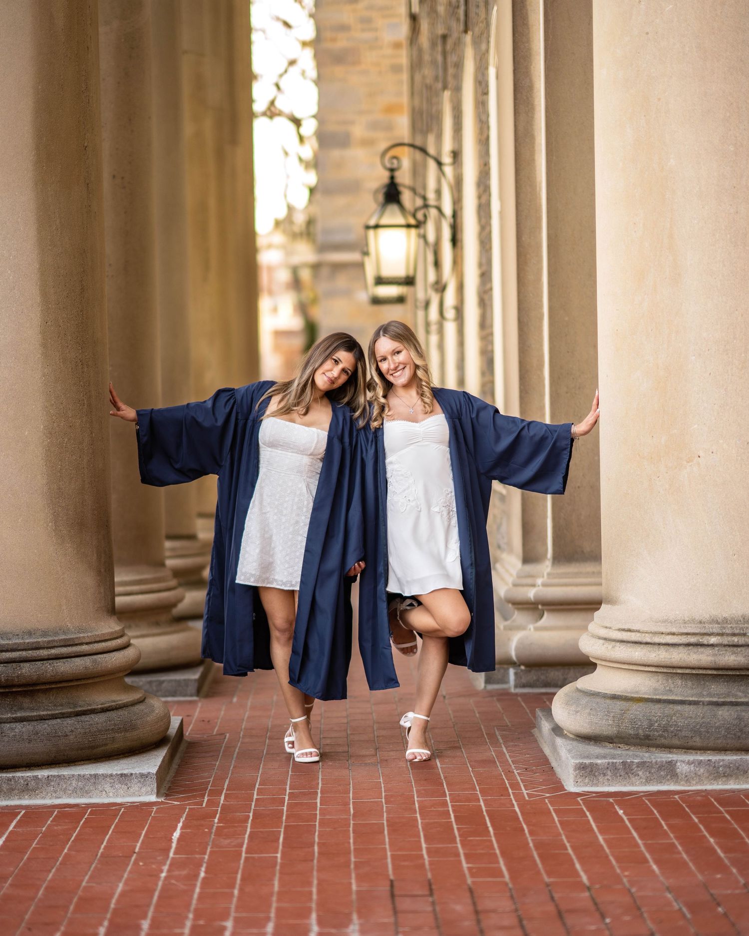 Two women in navy graduation gowns and white dresses pose playfully between columns on a brick walkway.