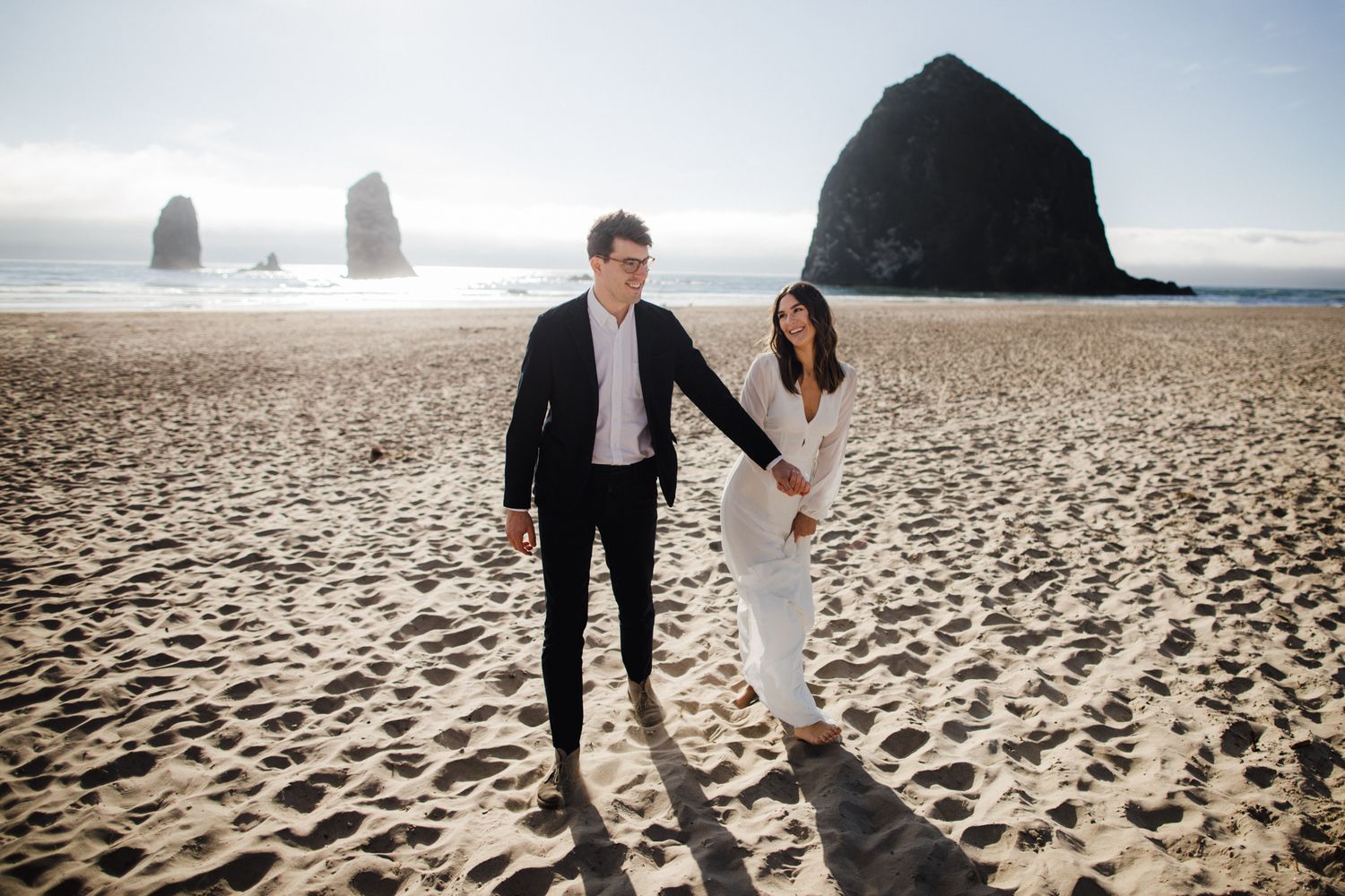Couple holding hands walking on Cannon Beach with Haystack Rock in the background on a sunny day.