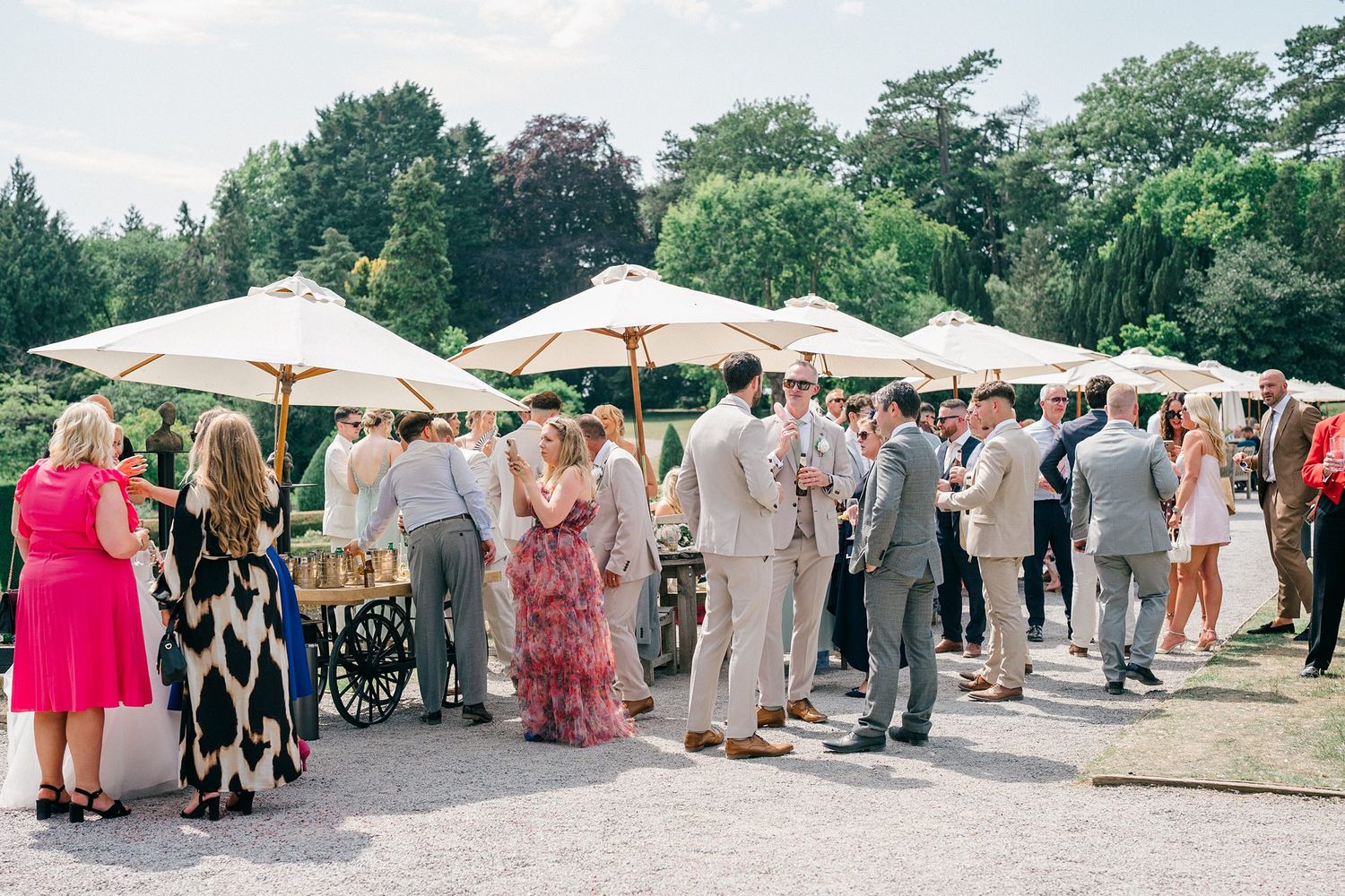 Elegant outdoor wedding reception with guests mingling under cream parasols in a lush garden setting.