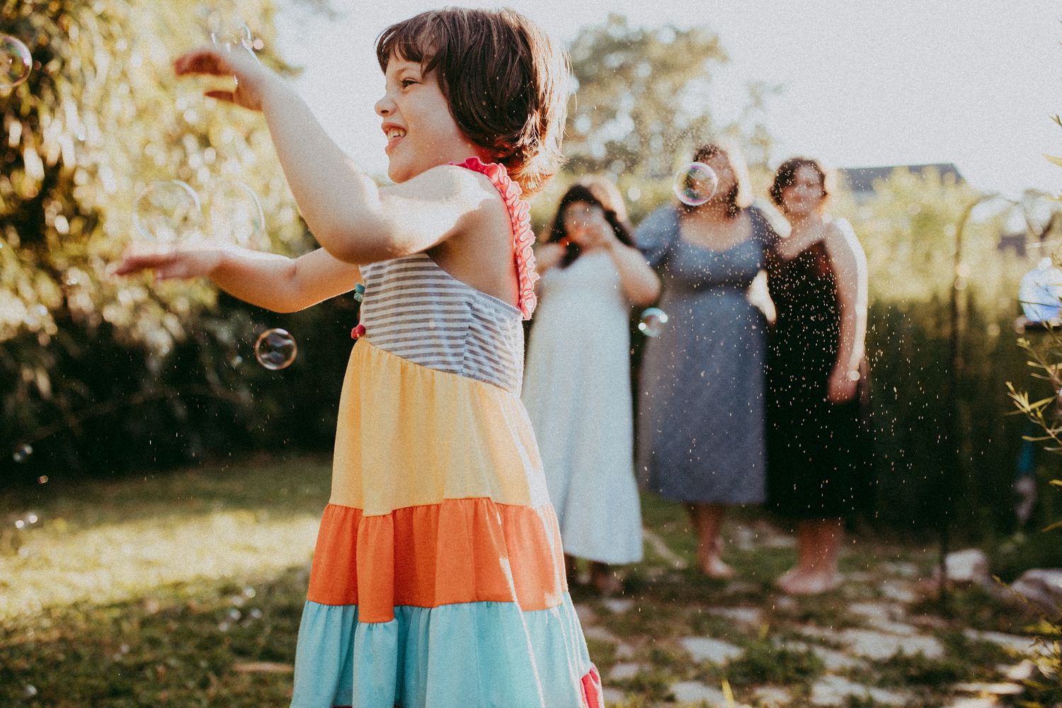 A young girl in a colorful tiered dress plays with bubbles outdoors while women in summer dresses socialize in the background. Whitney Popp