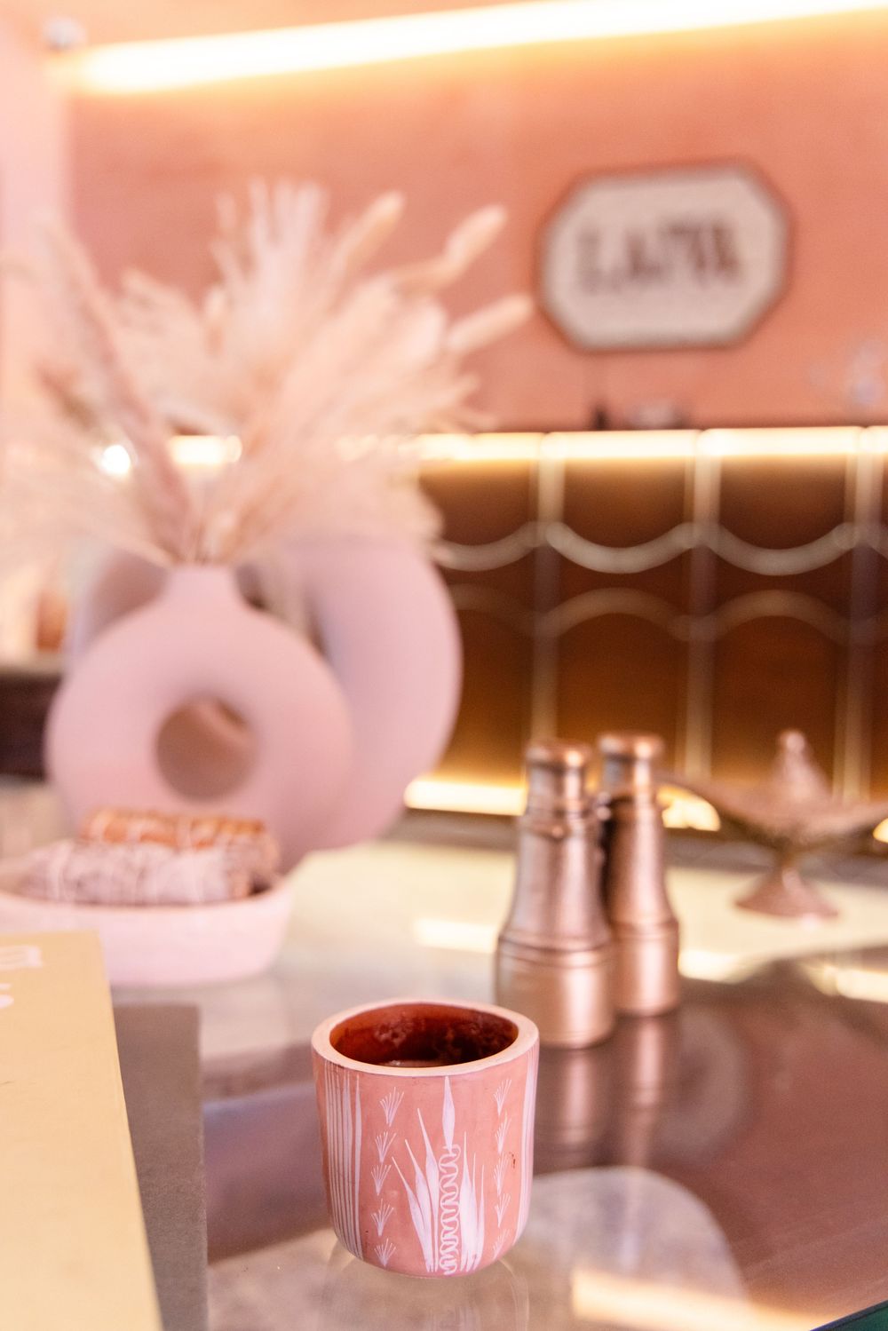 Pink ceramic cup on glass counter with donut-shaped vase and blurred cafe interior in warm lighting.