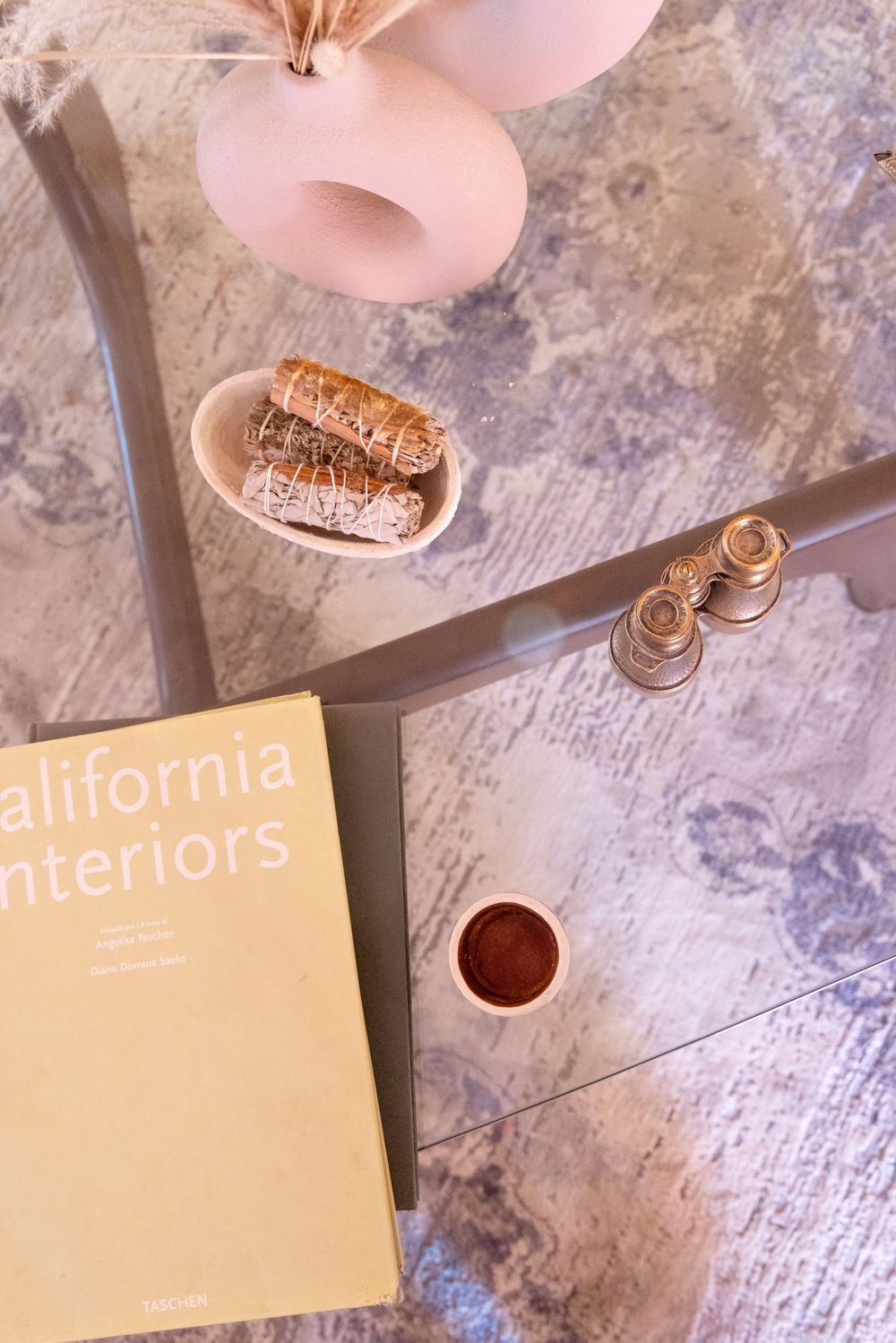 Overhead view of California Interiors book, sage bundles, pink vase, and gold candleholders on glass table.