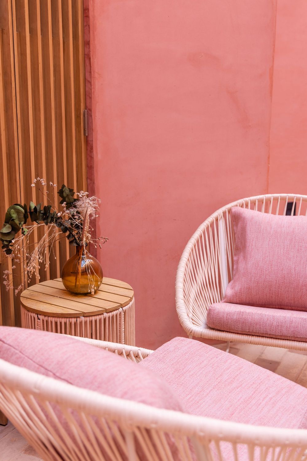 Pink rattan outdoor chairs with cushions beside a wooden side table with gold vase on pink patio.