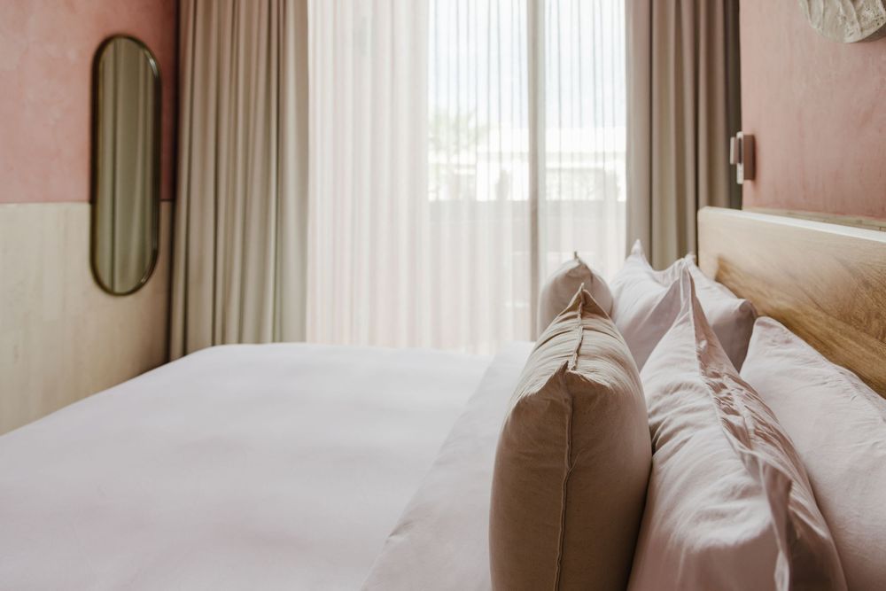 Cozy hotel bedroom with white bedding, blush pillows, wooden headboard, and sheer curtains by window.