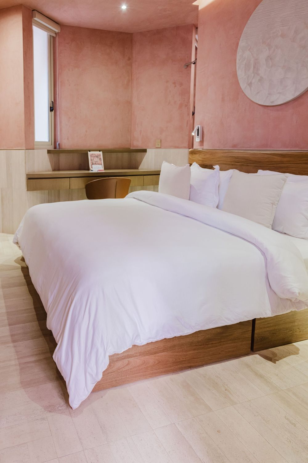 Bright hotel bedroom with white bedding, wooden bed frame, pink walls, and natural light from window.