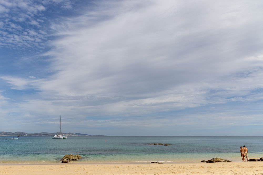 Serene sandy beach with calm turquoise water, anchored sailboat, and couple standing at shoreline.