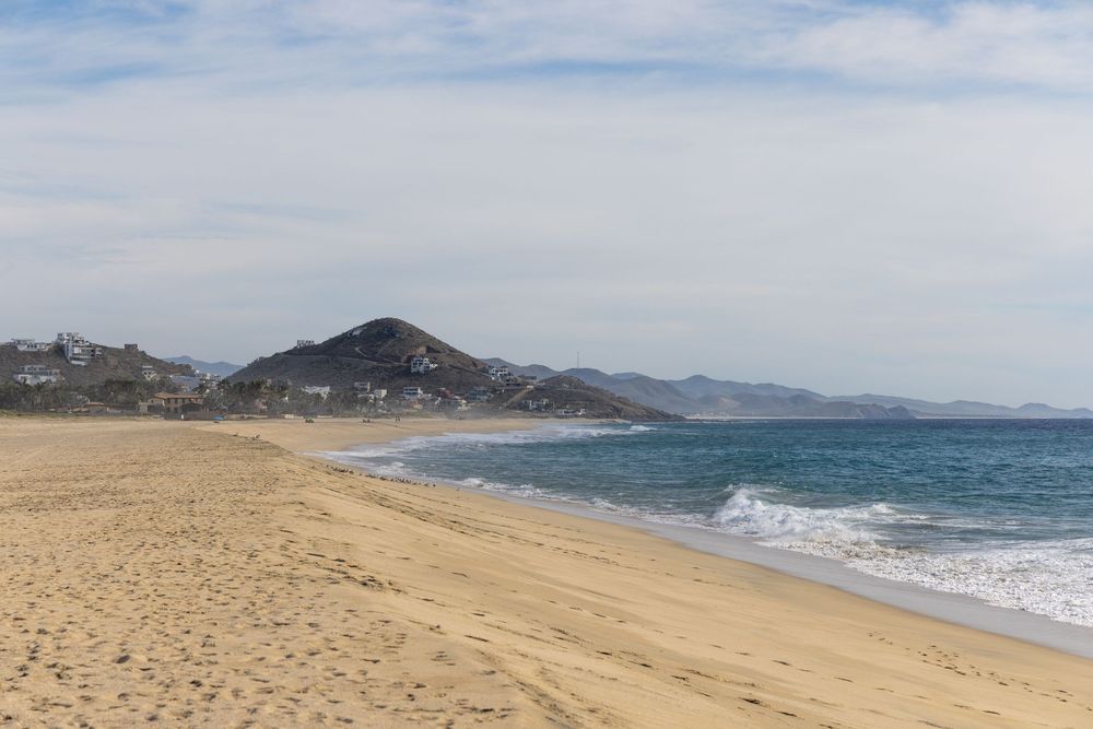 Scenic sandy beach with gentle ocean waves and a mountain in the background under cloudy skies.