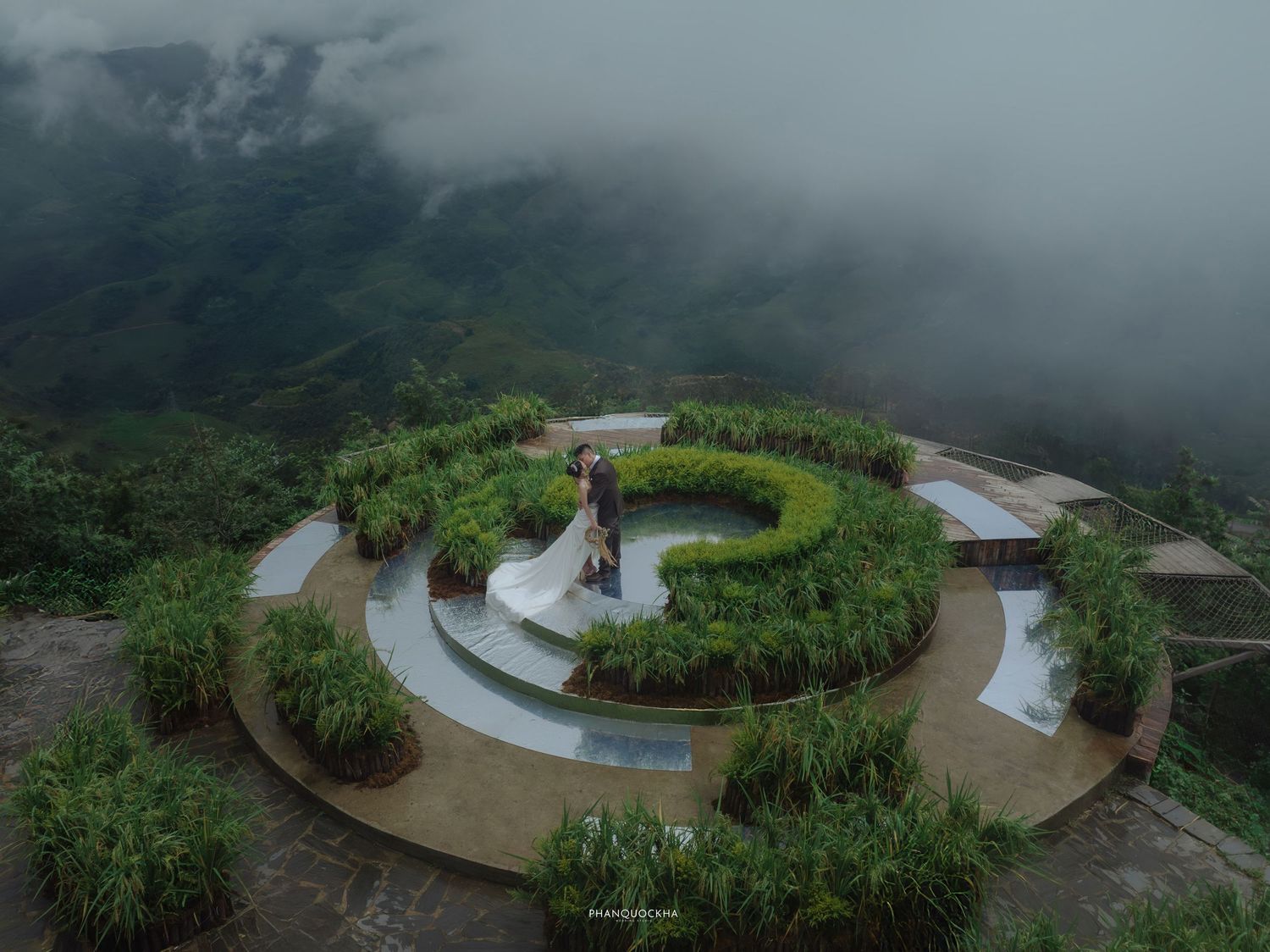 Aerial view of couple in wedding attire on circular hilltop garden surrounded by misty mountains.