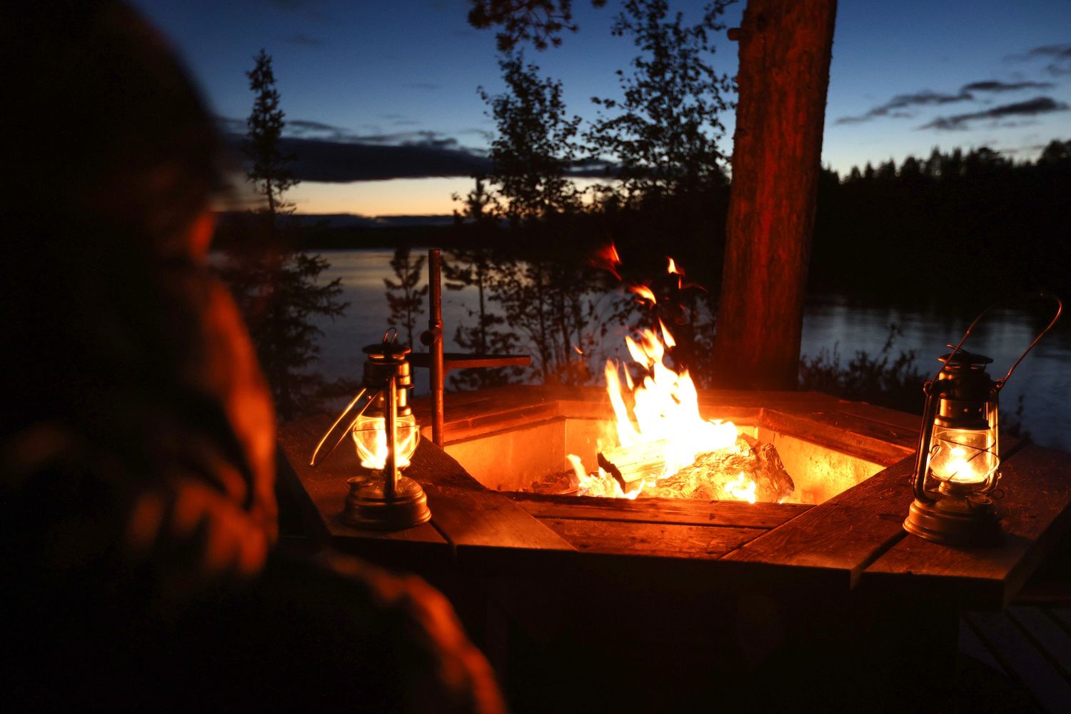Cozy campfire with lanterns beside a tranquil lake at twilight in the wilderness.