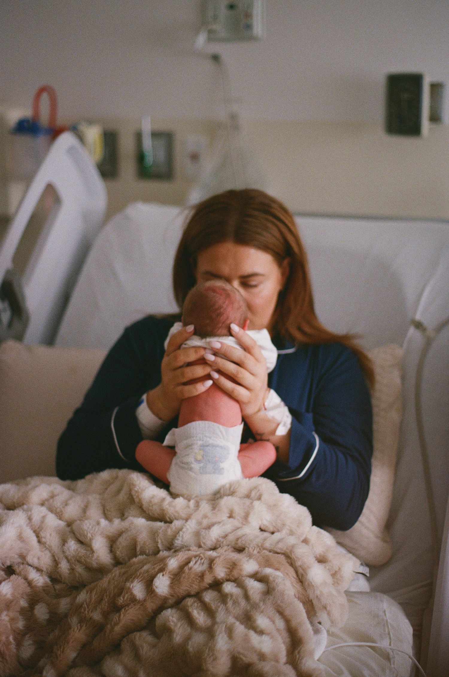 A new mother in a hospital bed tenderly kisses her newborn baby wrapped in a fluffy blanket.