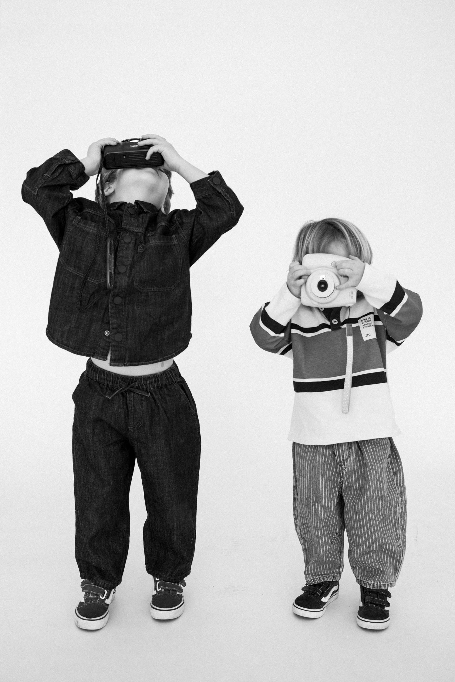 Two children pose with cameras in black and white, wearing casual denim and striped outfits with Vans sneakers.