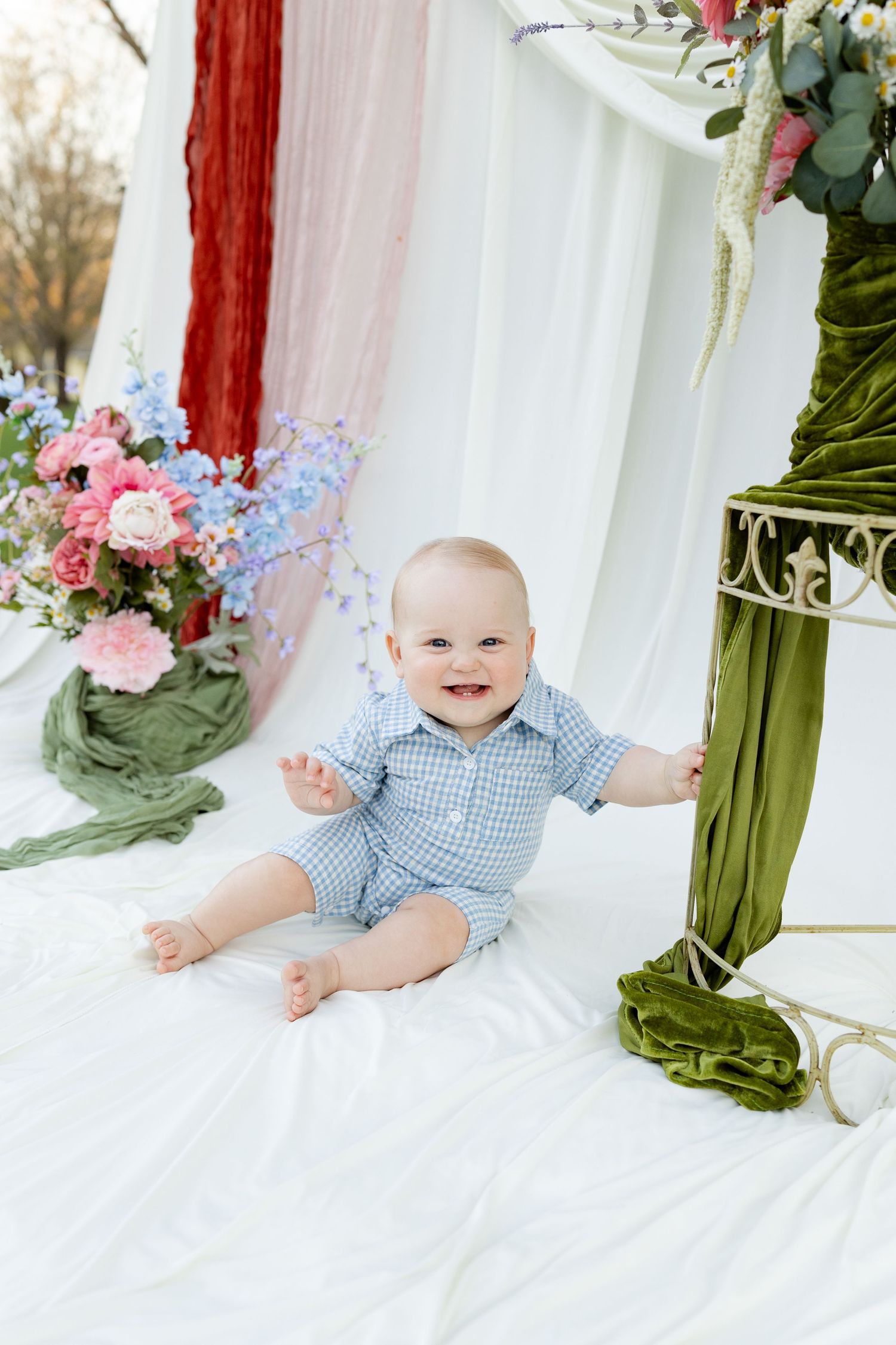 A smiling mother in a denim dress holds her happy baby boy in a floral studio portrait setting.