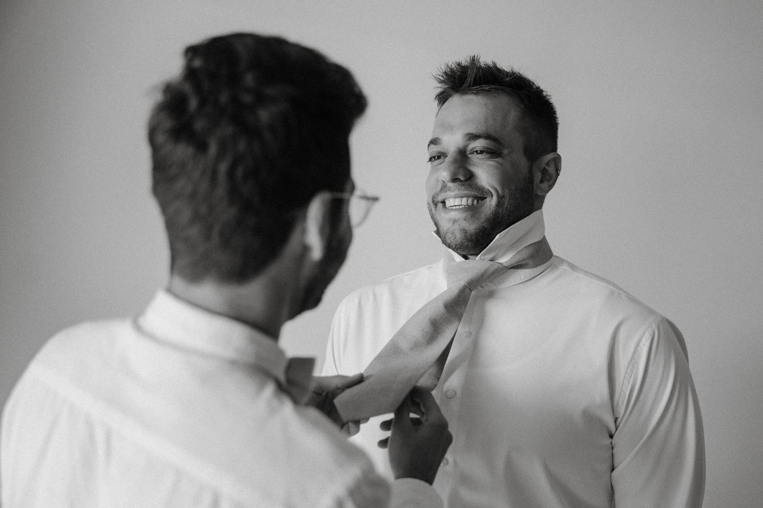 Black and white photo of a smiling groom having his tie adjusted by a groomsman before the wedding ceremony.