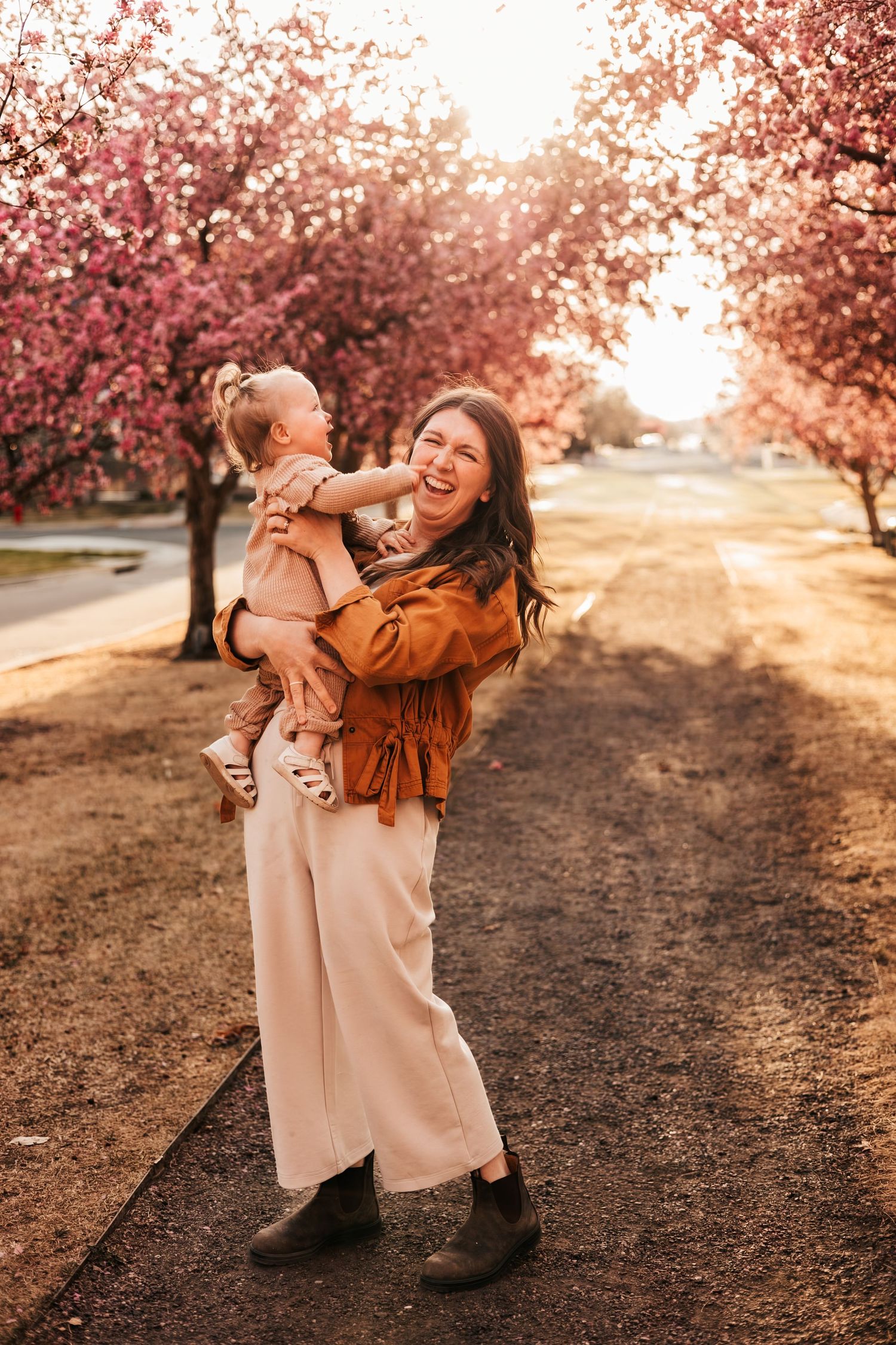 A laughing woman holds a toddler among pink cherry blossom trees on a sunlit path  | How to Get Nice Family Photos with a Wild Toddler