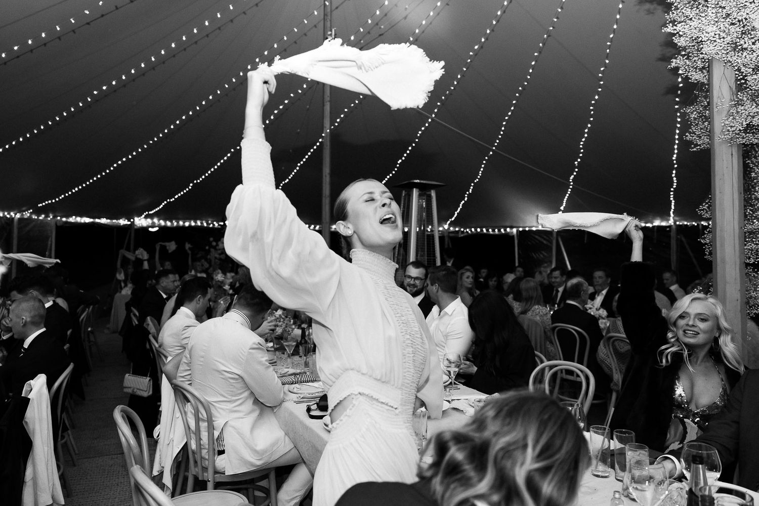 A joyful celebration at a wedding reception under a string light canopy with someone waving a white napkin in the air.
