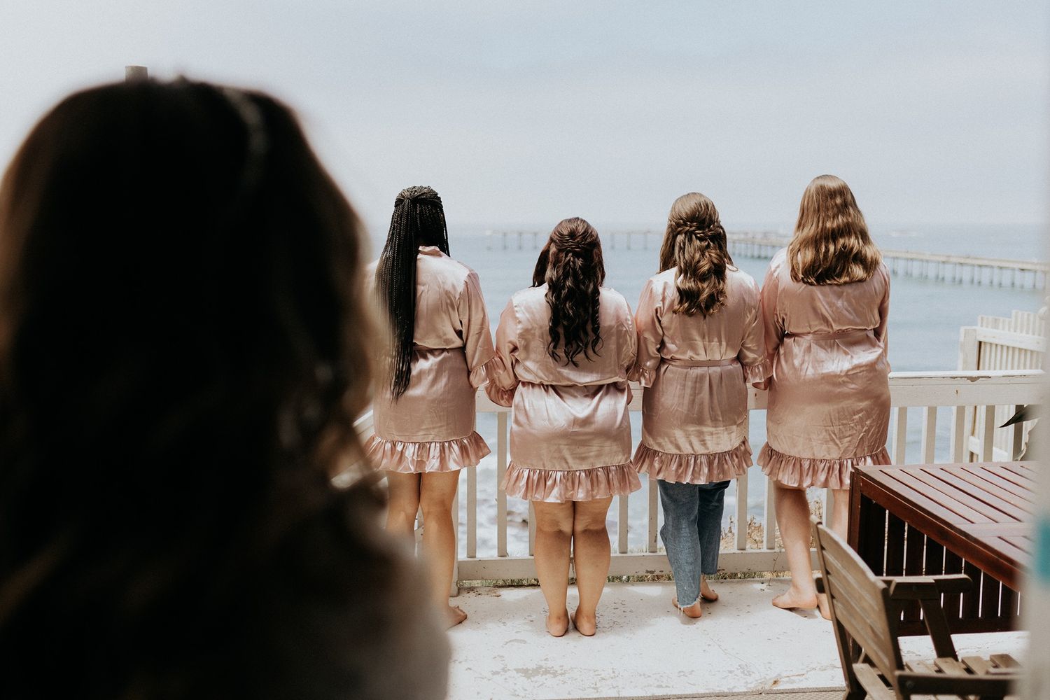Four bridesmaids in matching pink satin robes stand at a railing overlooking the ocean on a wedding day.