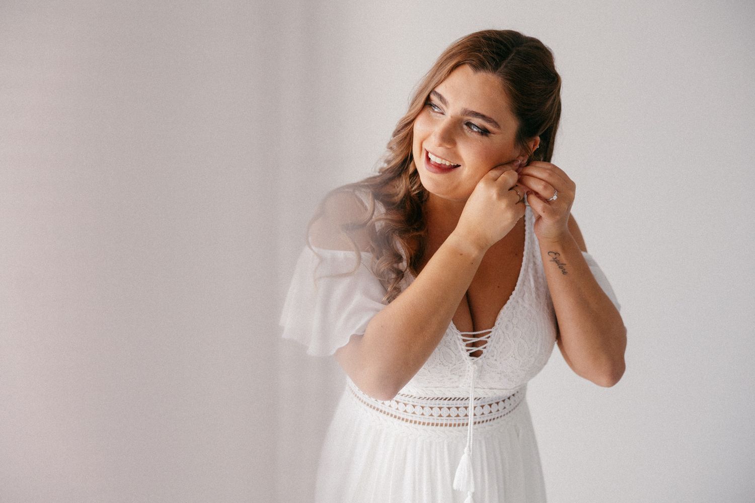 Plus-size bride in white lace dress putting on earrings, smiling elegantly against a light background.