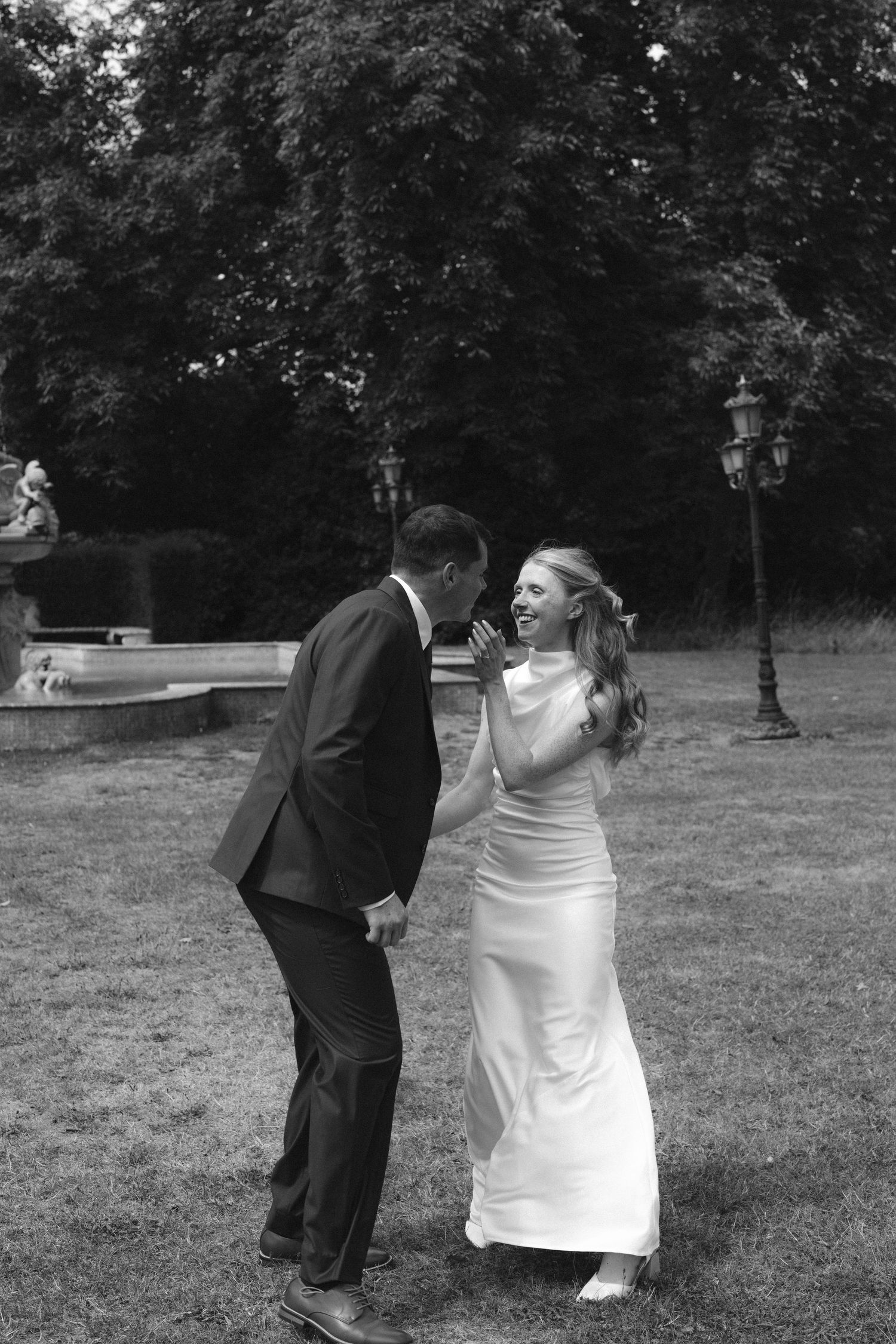 Black and white photo of a laughing bride in white gown and groom in suit sharing a joyful moment in a garden.