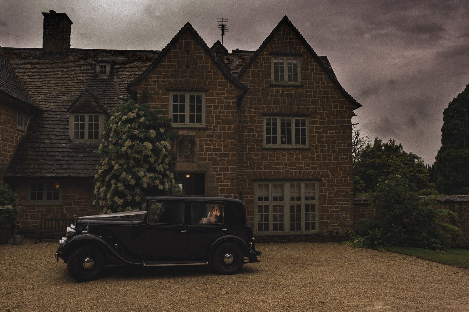 Vintage black car parked on gravel drive before a grand stone manor house under dramatic stormy skies.