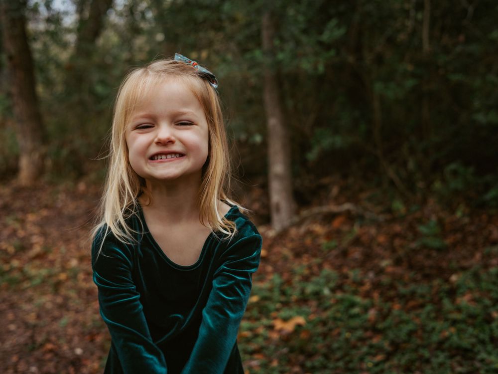 A candid lifestyle portrait of a smiling young girl in a green velvet dress during a natural light motherhood session in a wooded area near
