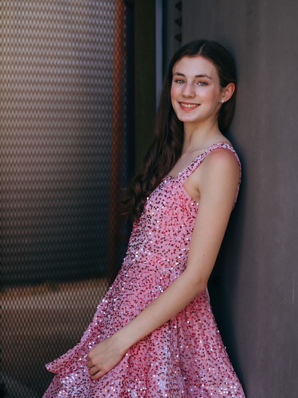 An editorial lifestyle portrait of a young woman in a pink dress leaning against a dark wall, showcasing refined lighting and a natural pose
