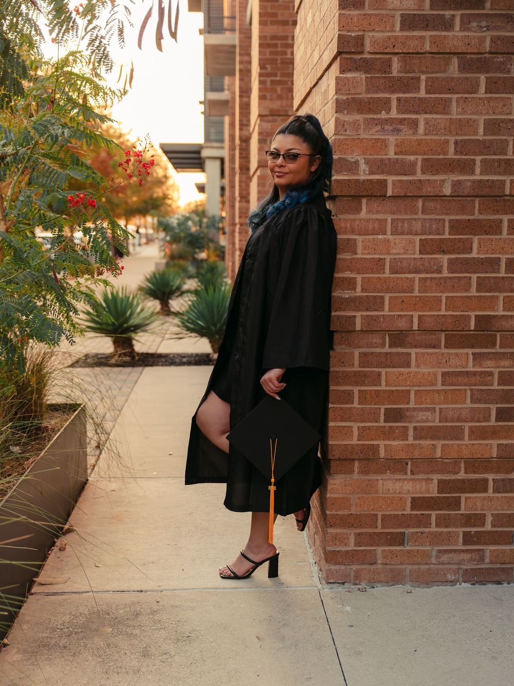 An editorial graduation portrait of a student in a black gown posing against a brick wall, showcasing a refined and modern aesthetic.