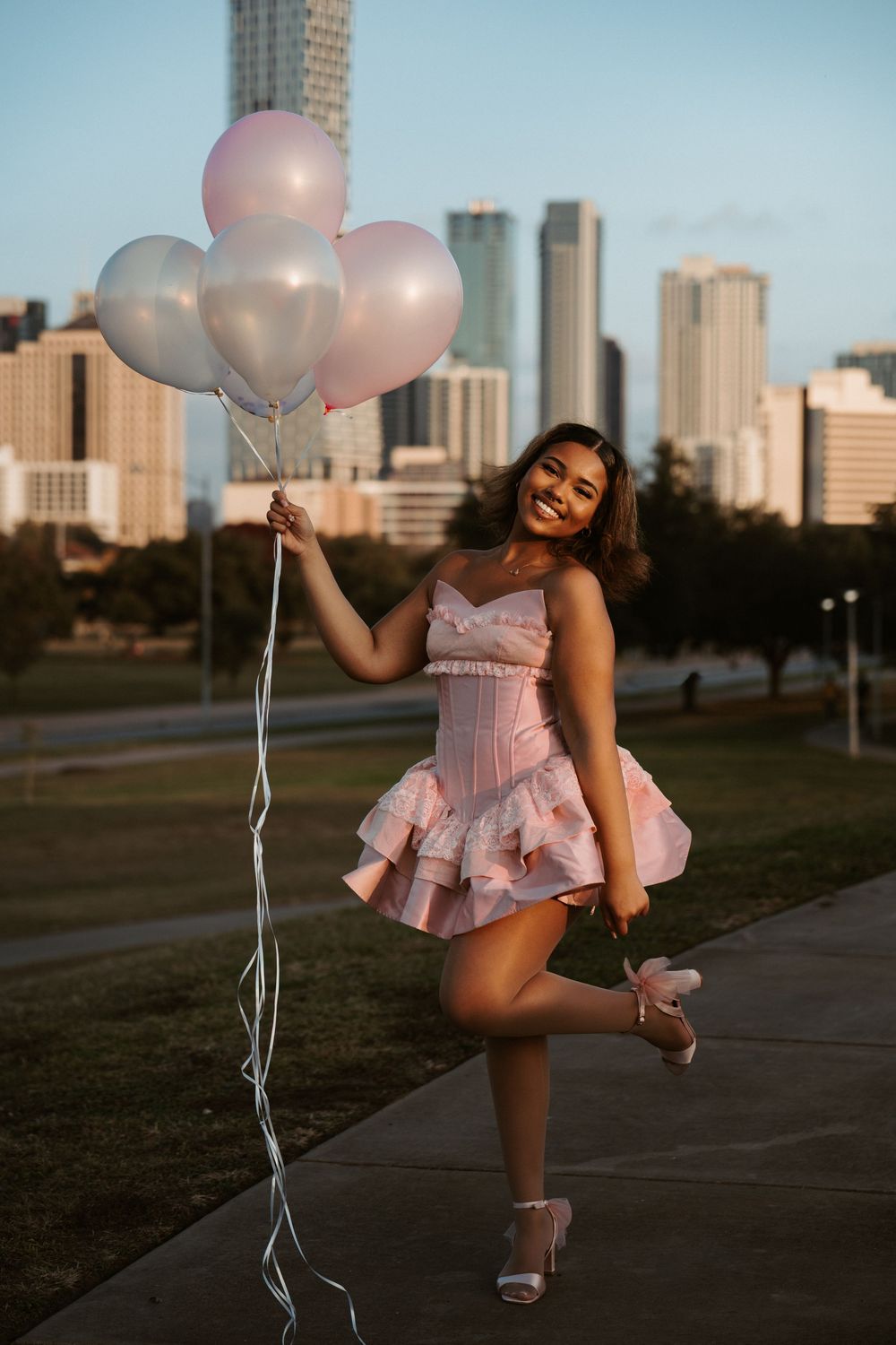 A full-length lifestyle portrait of a woman in a pink dress holding balloons with the Austin, Texas skyline in the background.