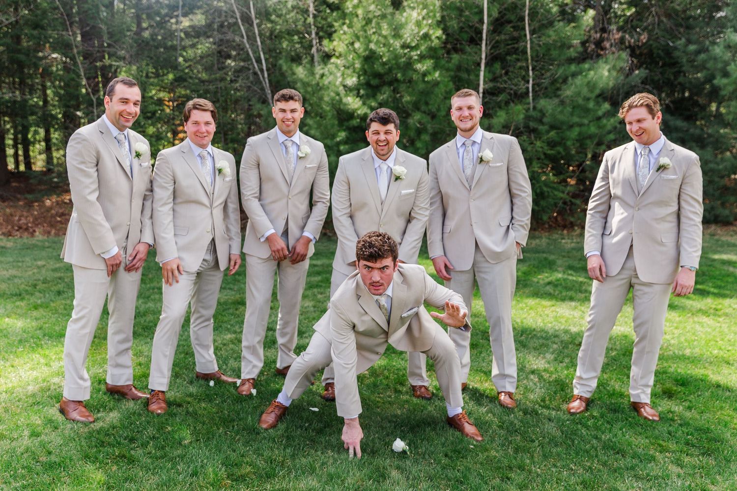Groomsmen in tan suits pose outdoors on grass, with one man doing a fun pose in front of the group at a wedding.