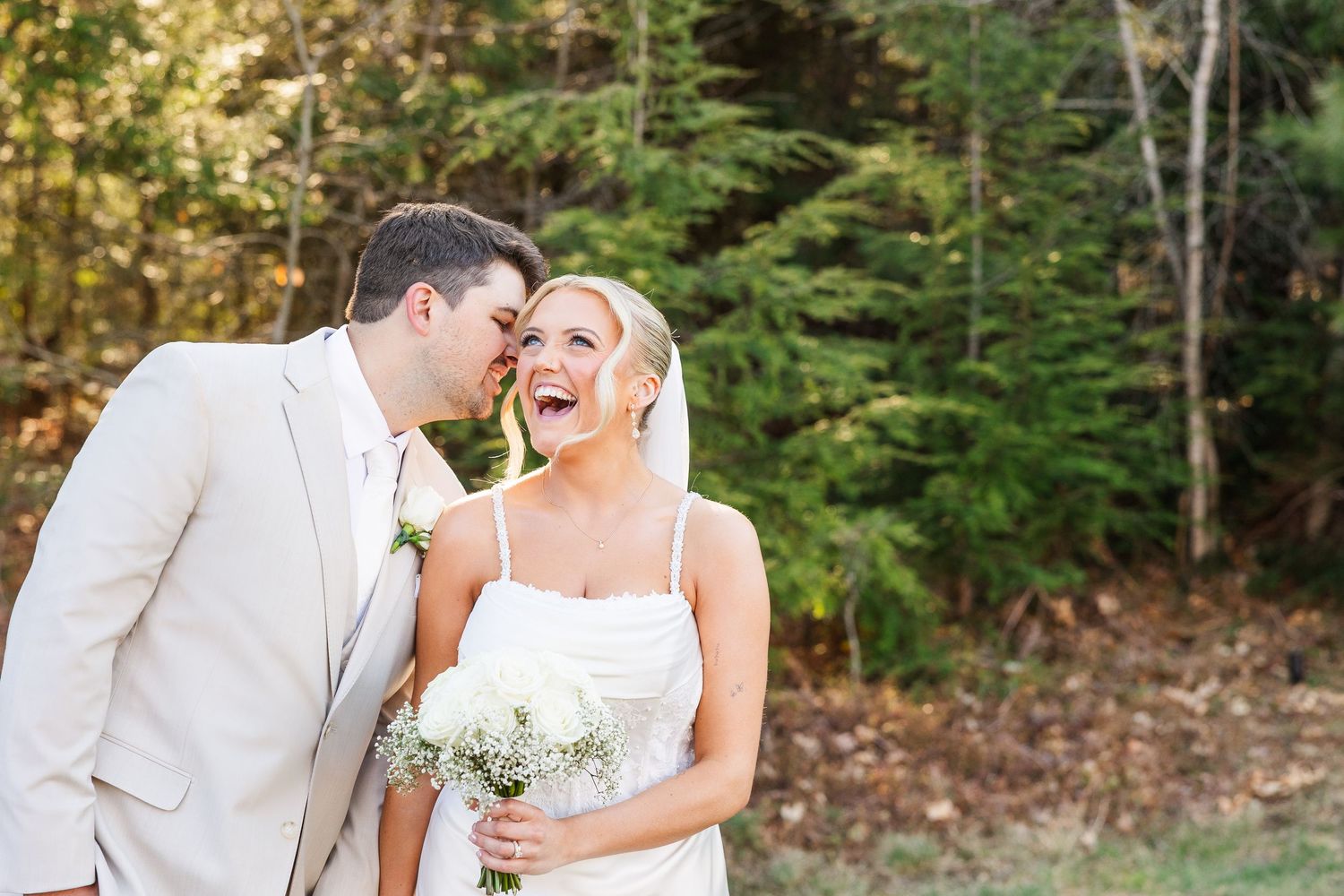 A laughing bride holding a white bouquet stands with a groom in a tan suit outdoors near evergreen trees.