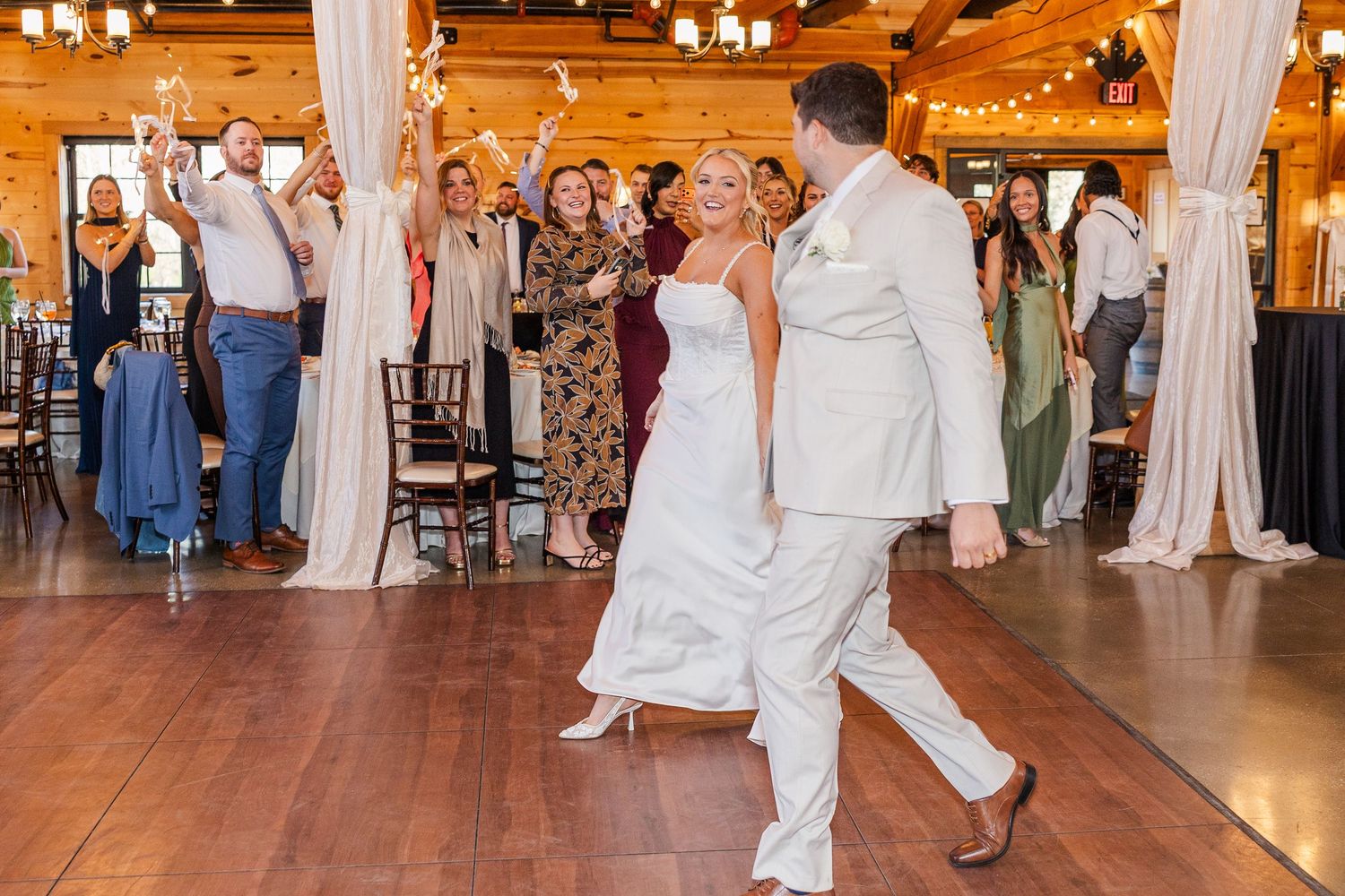 Bride and groom share first dance at rustic wedding reception while guests cheer and celebrate in elegant wood-paneled venue.