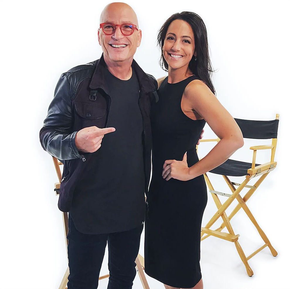 A man in a gray suit and woman in teal top smile together on a TV set with director chairs behind them.
