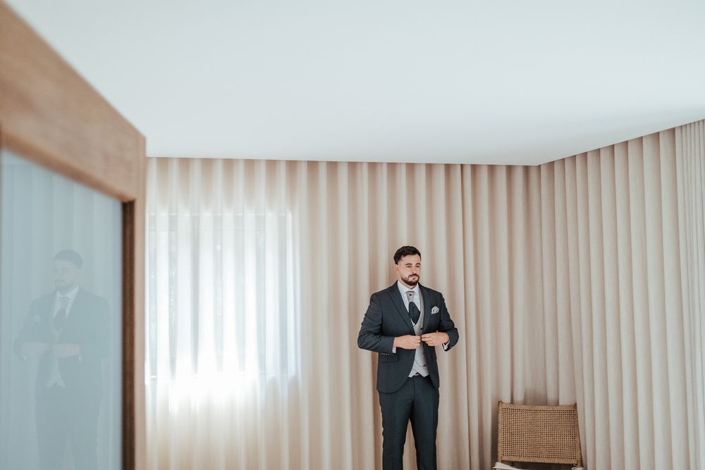 Groom in a dark suit adjusting his cufflinks while standing in an elegant room with beige curtains.