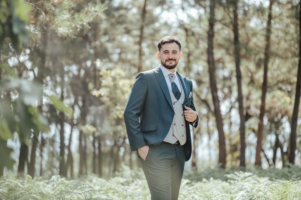 Smiling groom in dark suit and vest poses outdoors among lush green trees in a forest setting.