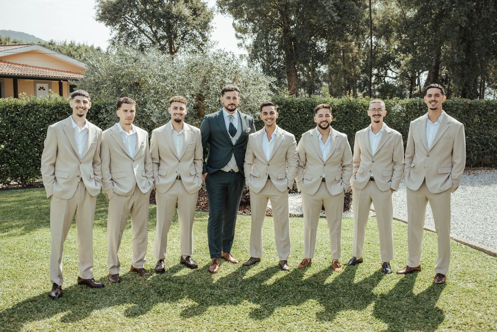 Groom in navy suit stands with eight groomsmen in matching tan suits outdoors on green lawn at wedding.