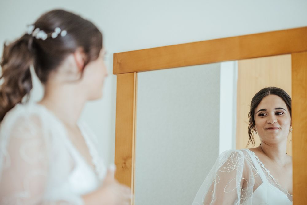 Bride in white lace robe admiring her wedding look in a wooden-framed mirror during bridal preparations.