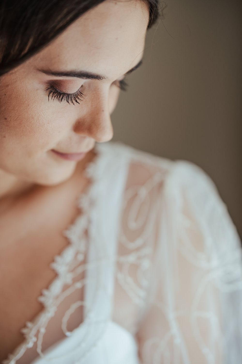 A bride in a white lace gown looks down serenely, showcasing elegant eye makeup and long lashes.