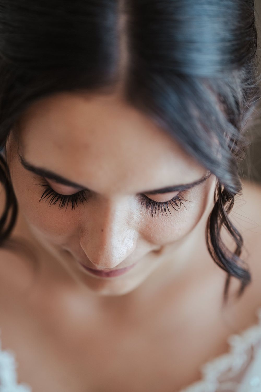 Close-up portrait of a bride with freckles, dark hair, and long lashes, looking down with a soft smile.