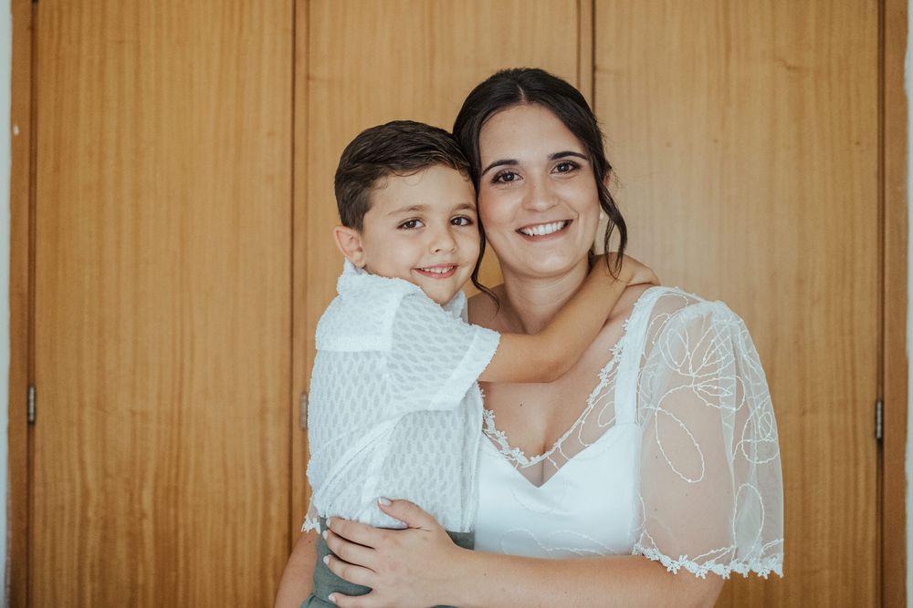 A smiling woman in a white lace dress hugs a young boy in white attire, standing in front of wooden doors.