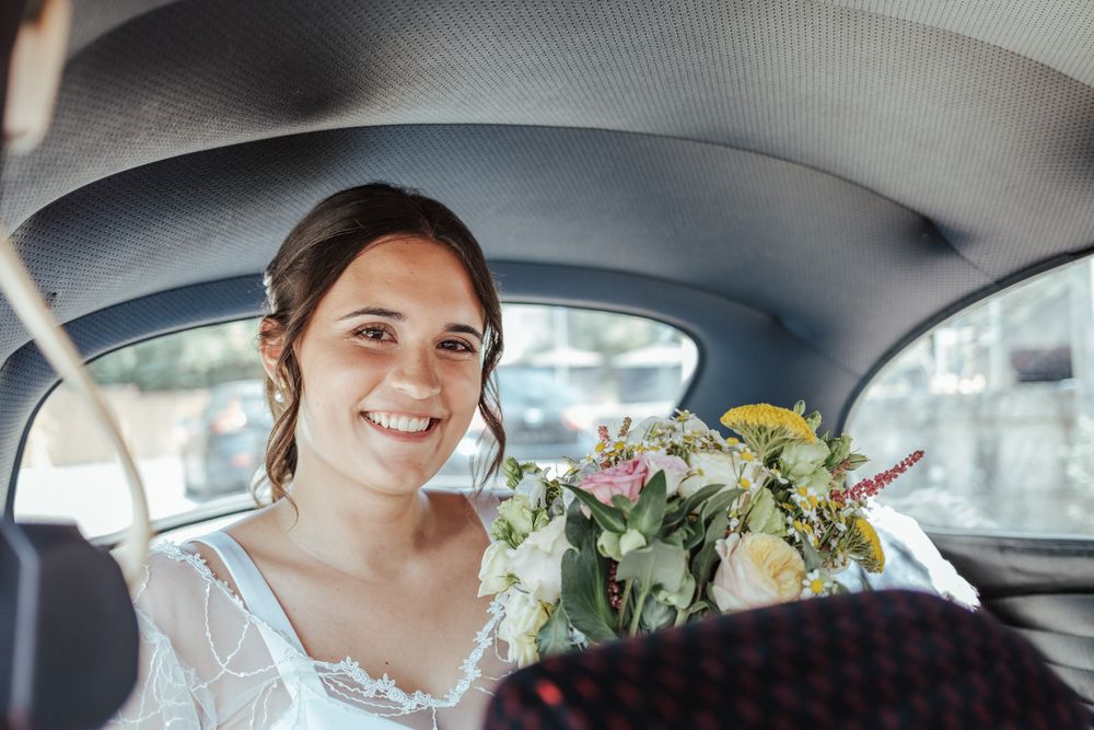 Smiling bride holding a colorful bouquet of flowers while sitting in the backseat of a vintage car on her wedding day.