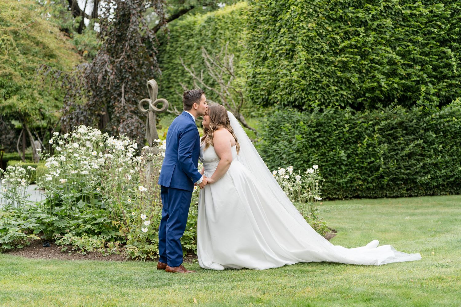 A bride and groom share a romantic kiss in a lush garden, with the bride in a flowing white gown and groom in a blue suit.