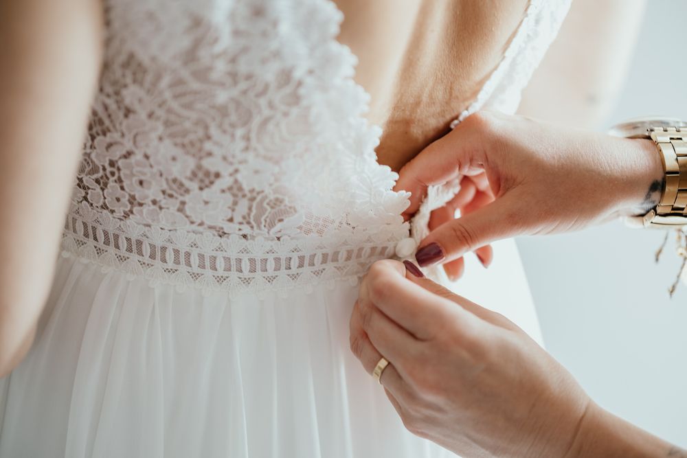 Close-up of hands fastening buttons on a white lace wedding dress from behind.