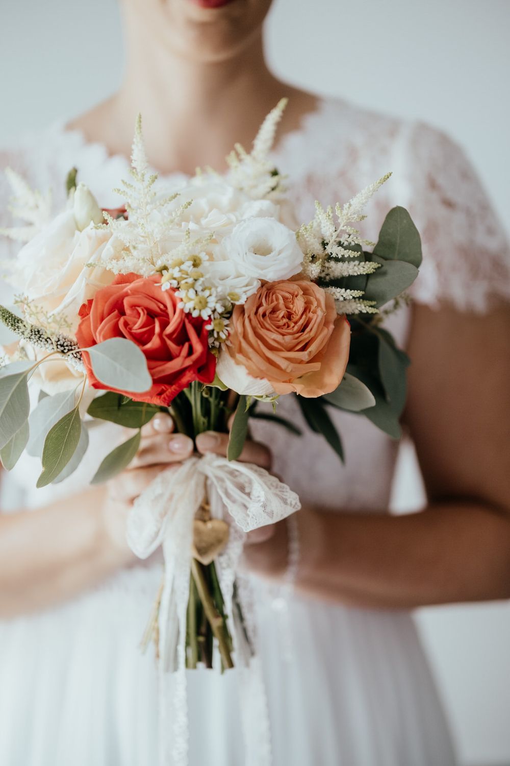 Bride in white lace dress holding a bouquet of coral, peach roses, white flowers, and eucalyptus greenery.