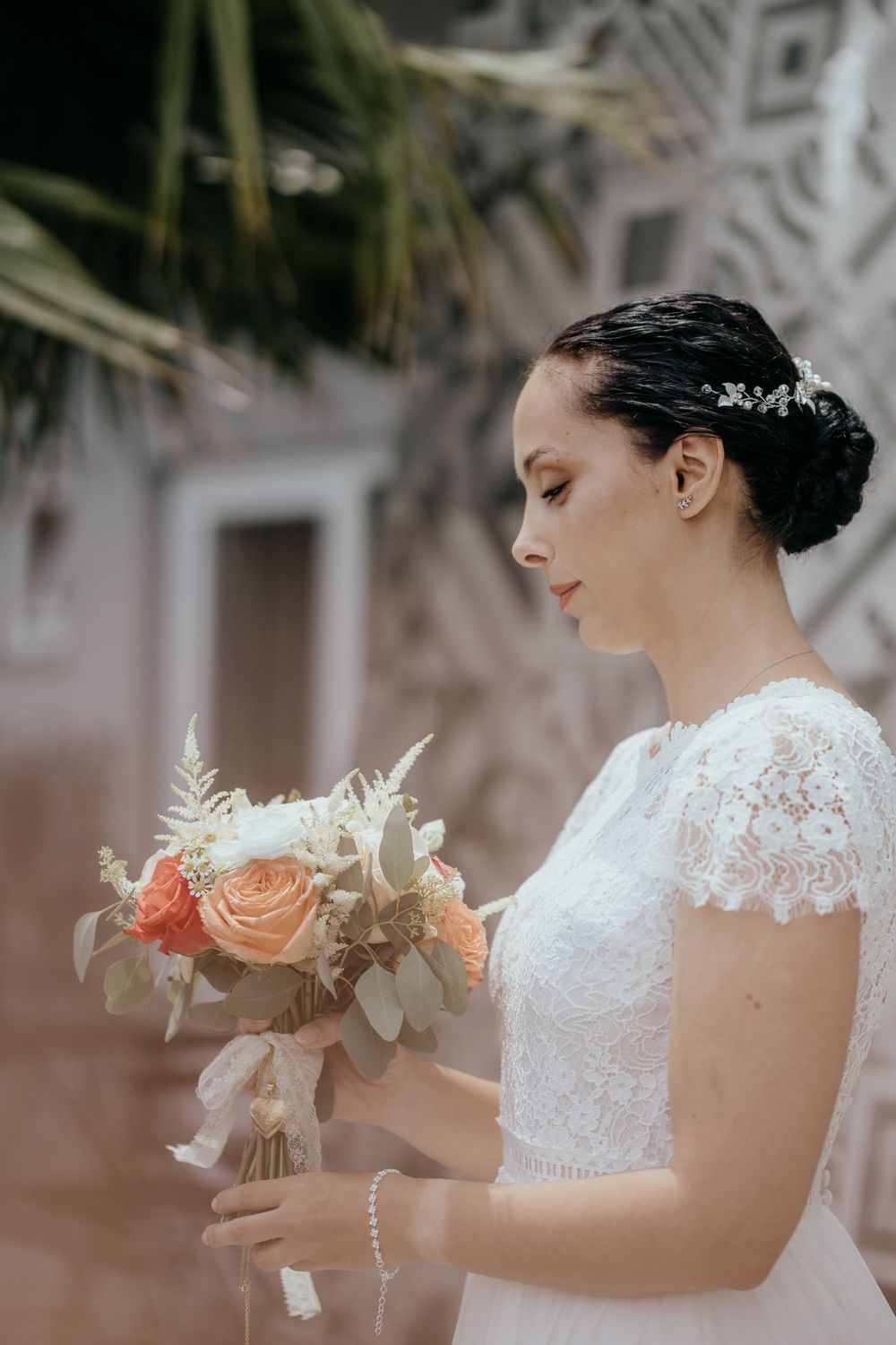 Elegant bride in lace wedding dress holding a bouquet of peach roses and white flowers, looking down gracefully.