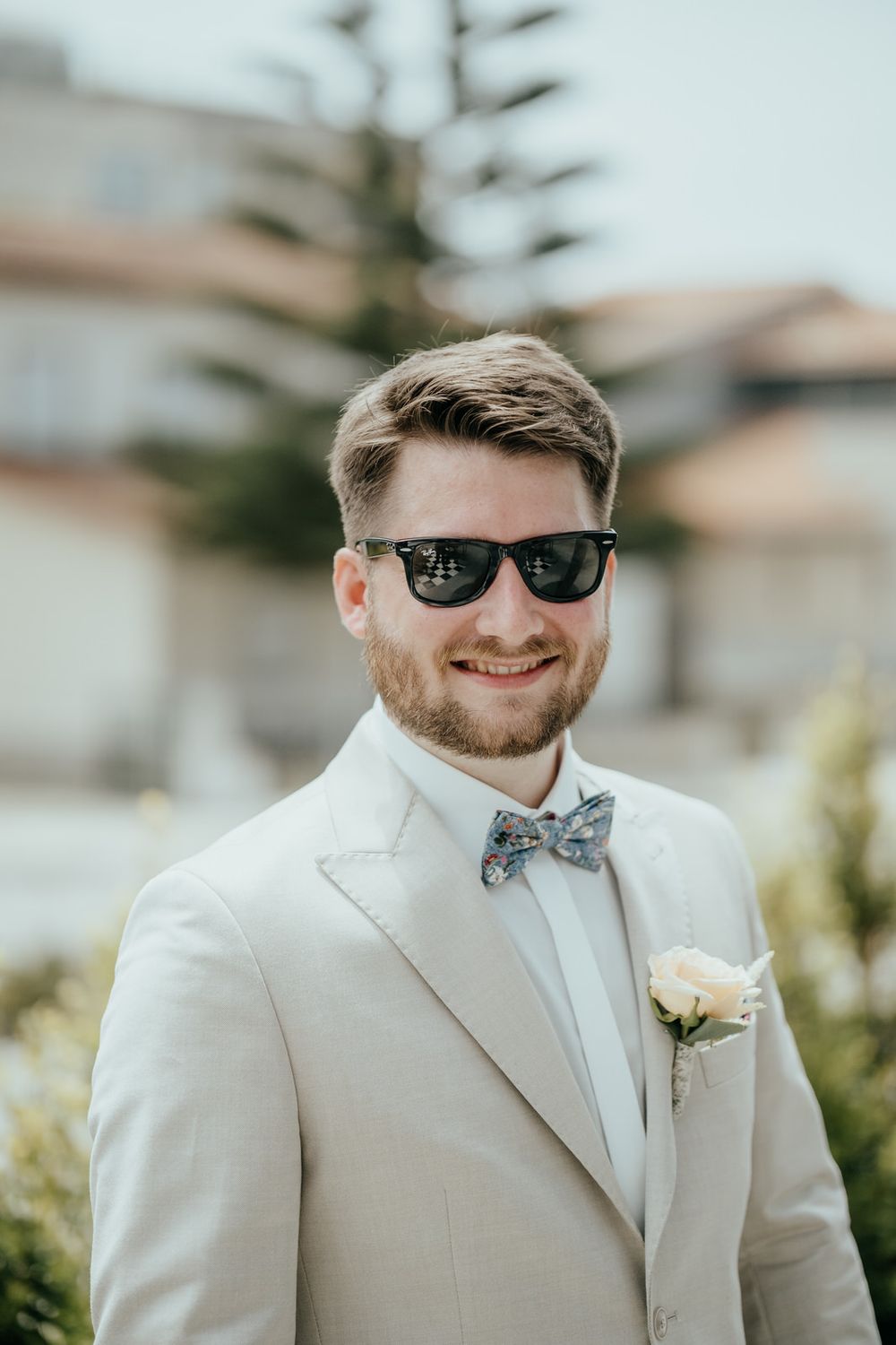 Smiling groom in white suit, floral bow tie, boutonniere, and sunglasses posing outdoors on his wedding day.