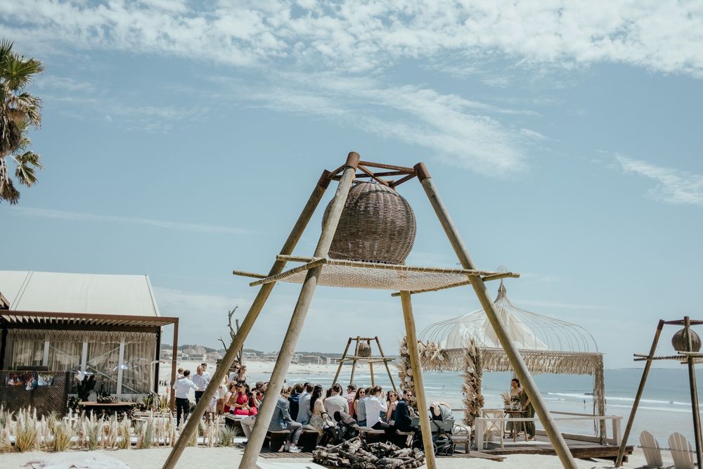 Rustic wooden tripod with wicker basket centerpiece at a beachside wedding ceremony under blue skies.